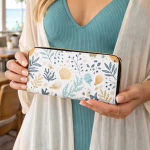 Puede incluir: Una cartera blanca rectangular con cremallera negra y detalles dorados. La cartera presenta un estampado botánico y de conchas marinas en tonos azules, dorados y verdes. Parece estar hecha de un material duradero.