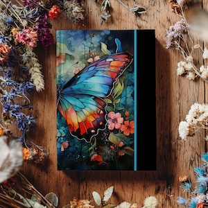 May include: A journal with a colorful butterfly design. The butterfly has blue, orange, and red wings, set against a backdrop of flowers and foliage. The journal has a black spine and is surrounded by dried flowers on a wooden surface.
