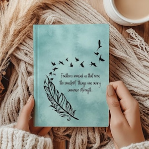 May include: A teal journal with a feather and birds design. The journal cover has the quote, "Feathers remind us that even the smallest things can carry immense strength." The journal is held by a person wearing a cream-colored sweater.