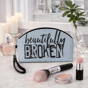 Könnte beinhalten: Hellblaue Kosmetiktasche mit dem schwarzen Schriftzug "beautifully BROKEN". Ein Make-up-Pinsel, Lippenstift und Rouge liegen neben der Tasche. Parfüm und Kerzen sind im Hintergrund.