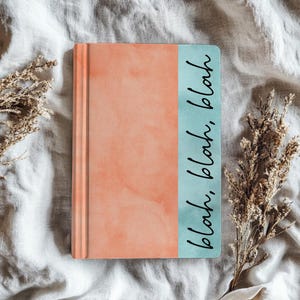 May include: A rectangular journal with a coral-colored cover and a teal spine. The spine features the words "blah, blah, blah" in a handwritten style. The journal is lying on a white, textured surface, with dried plants on either side.