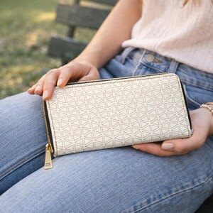 May include: Cream-colored wallet with a geometric triangle pattern. The wallet has a black zipper and gold-colored zipper pull. The wallet is rectangular and appears to be made of a leather-like material.