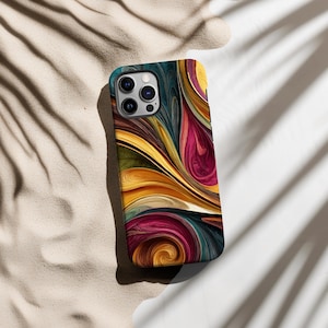 May include: A smartphone case with a colorful abstract design. The case features a swirling pattern of yellow, orange, pink, teal, and dark blue. The case has a camera cutout and is resting on a sandy surface.