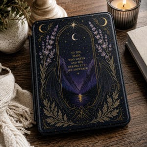 Puede incluir: Una funda para lector electrónico negra con una ilustración dorada de ramas de lavanda, lunas crecientes y alas que enmarcan un arco central. El texto dice: TO THE STARS WHO LISTEN AND THE DREAMS THAT ARE ANSWERED, sobre un paisaje montañoso.