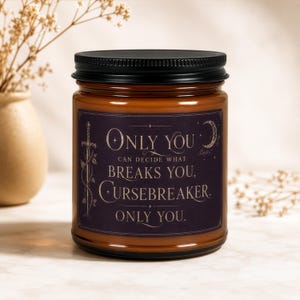 Op de afbeelding: Een bruine kaars in een glazen pot met een zwarte deksel. Het etiket is donkerpaars met een zwaardillustratie, een maansikkel en de tekst: Only you can decide what breaks you, Cursebreaker. Only you.