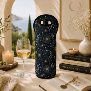 Peut inclure: Un refroidisseur de bouteille de vin bleu marine avec un motif céleste avec des étoiles, des lunes et des soleils dorés. Le refroidisseur est posé sur une table à côté d'un verre de vin blanc, d'une bougie allumée et d'un livre.