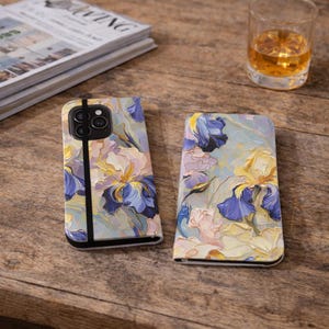 May include: Two phone cases with a floral design featuring blue, yellow, and pink irises. One case is open, revealing the phone's camera, while the other is closed. The cases rest on a wooden surface, with a glass of amber liquid and a magazine in the background.