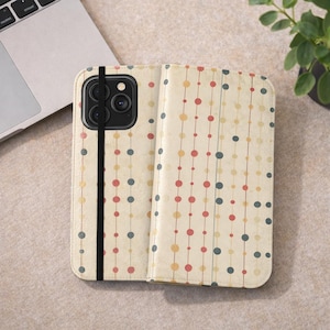 Puede incluir: Una funda de teléfono beige con un patrón de puntos de colores. La funda está abierta, revelando la cámara del teléfono y una correa elástica negra. Los puntos son rojos, amarillos y azules, dispuestos en líneas verticales.