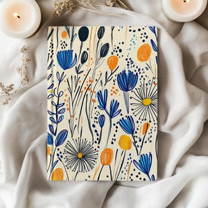 May include: A hardcover journal with a cream-colored background and a floral pattern. The design features blue, orange, and black stylized flowers and foliage. The journal is rectangular and appears to be closed.