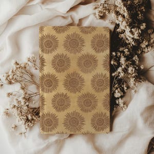 May include: A rectangular notebook with a repeating sunflower pattern in shades of brown on a tan background. The notebook is surrounded by white flowers and a white cloth.