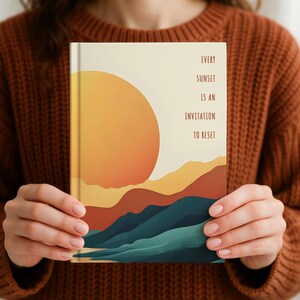 May include: A journal with a sunset-themed illustration. The cover features a large orange sun, layered mountains in shades of brown and blue, and the text "EVERY SUNSET IS AN INVITATION TO RESET". The journal is held by a person wearing a brown sweater.