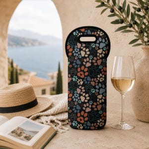 Peut inclure: Housse de bouteille de vin noire avec un motif coloré de pattes de chien dans des tons orange, bleu et rose. Le sac a une poignée noire et est fait d'un matériau semblable au néoprène. Un verre de vin avec du vin blanc est à côté du sac.