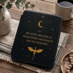 May include: A black e-reader case featuring a gold crescent moon, stars, and a dragon graphic. The text reads: BECAUSE ADULTING IS EXHAUSTING AND REALITY HAS NO DRAGONS. The case has a textured finish and is rectangular with rounded corners.