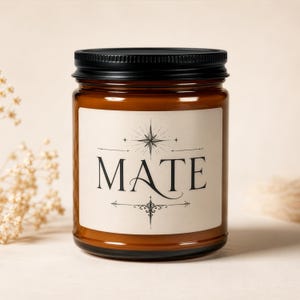 Pode incluir: Um pote cilíndrico de vidro âmbar para velas com tampa preta de rosca. O rótulo cor de creme na frente apresenta a palavra MATE impressa em fonte serifada preta, acentuada por um gráfico de estrela acima e um ornamento decorativo abaixo.