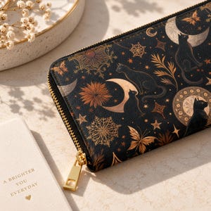 Könnte beinhalten: Ein schwarzes rechteckiges Portemonnaie mit goldenem Reißverschluss und einem himmlischen Muster aus schwarzen Katzen, Mondsicheln, Sternen, Schmetterlingen und komplexen floralen Motiven in Gold- und Kupfertönen auf einer strukturierten Oberfläche.