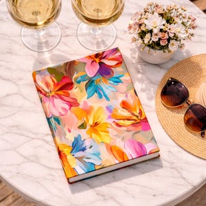 May include: A hardcover journal with a vibrant floral design in shades of pink, yellow, orange, and blue. The journal is on a marble table, next to a straw hat, sunglasses, and a small vase of flowers. Two glasses of white wine are also visible.
