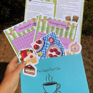 Könnte beinhalten: Ein hellblauer Umschlag mit der Aufschrift The Cuppa Post Club und einer Kaffeetassen-Illustration, der bedruckte Aktivitätsblätter, ein Wortsuchrätsel, eine Schnitzeljagd sowie dekorative Aufkleber von Erdbeeren, Kuchen und einer Schleife enthält.