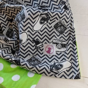 May include: Green and white fabric with a black and white chevron pattern and a repeating image of vintage cameras. The fabric has a white polka dot pattern on one side and a black and white chevron pattern with cameras on the other side. The fabric has a label that says "Little Wolf Garden".