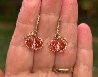 May include: A pair of gold-colored wire earrings, each featuring a spherical, faceted, reddish-orange crystal. The crystals are encased in a delicate wire cage, with a looped wire at the top for hanging. The earrings are displayed against a blurred green background.