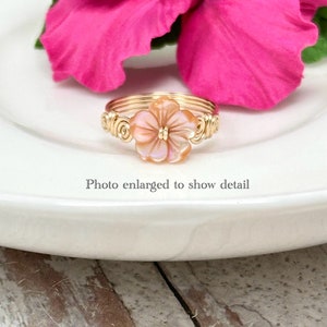 May include: A gold wire wrapped ring with a pink shell flower accent. The ring is on a white plate with a pink hibiscus flower in the background. "Photo enlarged to show detail" is written below the ring.