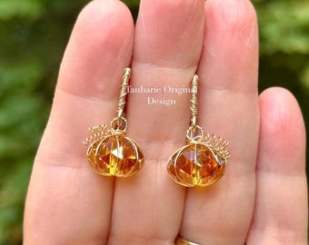May include: A pair of dangle earrings featuring faceted amber-colored crystal pumpkin charms wrapped in gold-toned wire. The earrings include coiled wire stems and small wire tendrils. The text Tanbarie Original Design is visible in the center.
