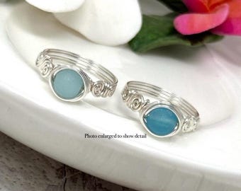 May include: Two silver wire rings with light blue oval stones. The rings have a spiral design on the sides. The rings are displayed on a white plate with a flower in the background. The text "Photo enlarged to show detail" is visible.