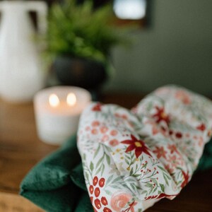 May include: A white fabric eye pillow with a floral pattern in shades of red, pink, and green. The pillow is folded and resting on a green fabric surface.