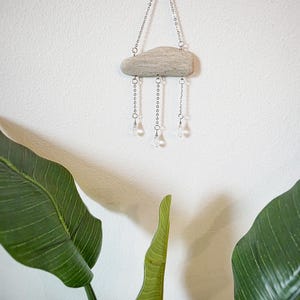 May include: A decorative wall hanging featuring a driftwood cloud shape suspended by a silver chain. Clear crystal beads hang below the cloud, adding a touch of sparkle. The piece is mounted on a white wall, with green leaves in the foreground.