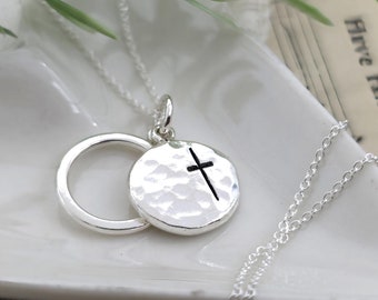 May include: A silver necklace with a hammered circle pendant featuring a small cross. The necklace is on a white surface.