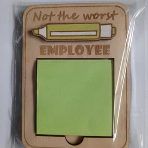 May include: Wooden memo board with the text "Not the worst EMPLOYEE" and a highlighter graphic. The board has a rectangular shape with rounded corners and a cutout for a green sticky note pad. The board is packaged in a clear plastic sleeve.