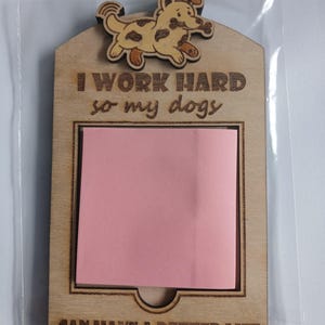 May include: Wooden memo board featuring a laser-cut dog design. The board is engraved with the text "I WORK HARD so my dogs CAN HAVE A BETTER LIFE" and includes a pink sticky note pad. It is packaged in a clear plastic sleeve.