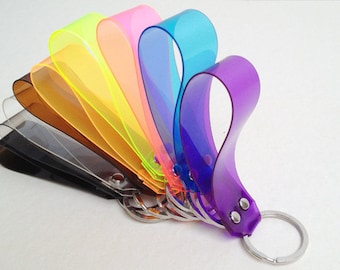 May include: A set of seven colorful keychains, each made of clear plastic with a different color. The keychains are arranged in a fan shape, with the purple keychain at the bottom and the yellow keychain at the top. Each keychain has a silver metal ring attached to it.