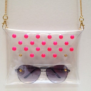 Puede incluir: Un bolso de pl&aacute;stico transparente con una correa de cadena dorada y lunares rosas. El bolso tiene un par de gafas de sol dentro.