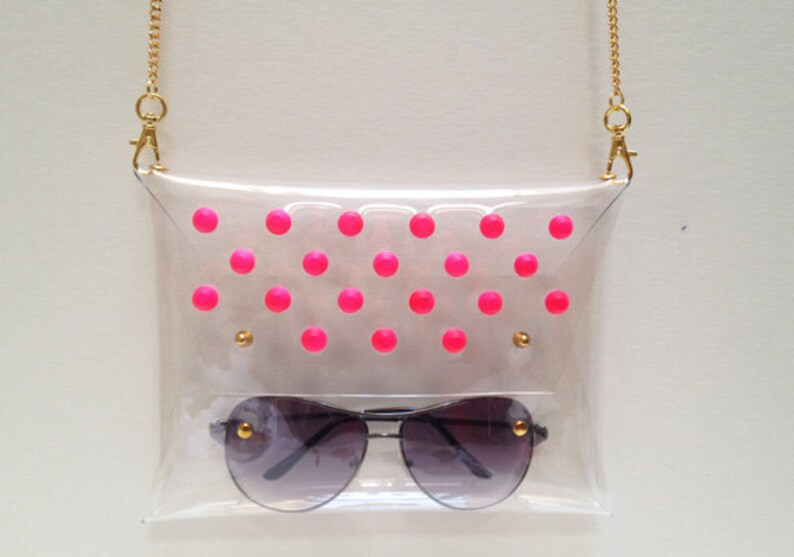 Puede incluir: Un bolso de pl&aacute;stico transparente con una correa de cadena dorada y lunares rosas. El bolso tiene un par de gafas de sol dentro.