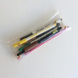 Puede incluir: Un estuche transparente que contiene varios bolígrafos y rotuladores. Los bolígrafos son de varios colores, incluyendo amarillo, verde, marrón, negro y rosa. Algunos bolígrafos tienen texto que dice "TURBO SCENT" y "Le Coloring Brush Pen".