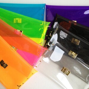 May include: A stack of seven clear plastic clutches in various bright colors, including orange, pink, green, blue, and purple. Each clutch has a gold clasp and gold studs around the edges.