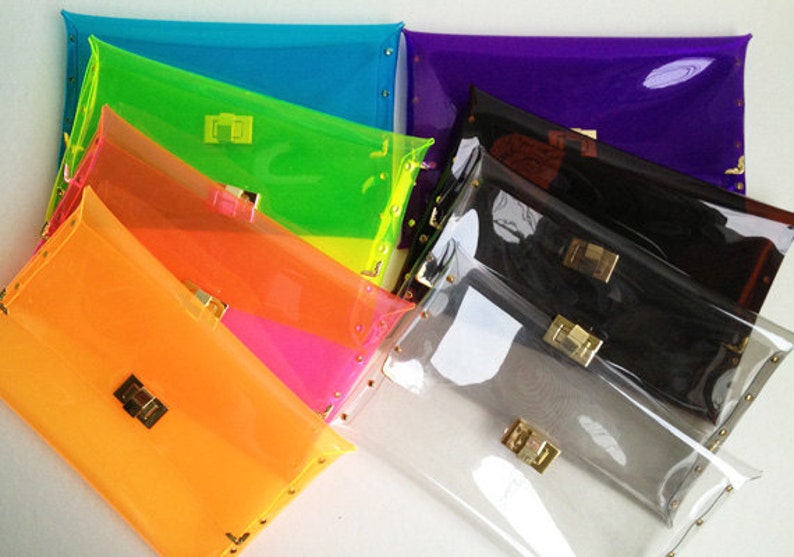 May include: A stack of seven clear plastic clutches in various bright colors, including orange, pink, green, blue, and purple. Each clutch has a gold clasp and gold studs around the edges.