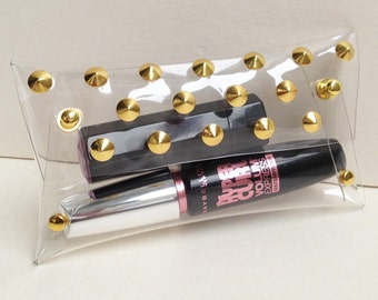 May include: Clear plastic makeup bag with gold pyramid-shaped studs. The bag contains a black mascara tube with the text "Maybelline The Falsies Super Curl Volume Express Waterproof" and a black and silver lipstick tube.