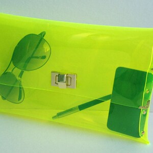 May include: A bright yellow transparent clutch bag with a gold clasp. The bag is open and contains sunglasses, a pen, and a green phone.