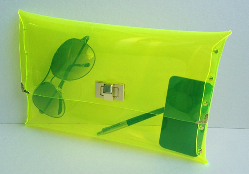 May include: A bright yellow transparent clutch bag with a gold clasp. The bag is open and contains sunglasses, a pen, and a green phone.