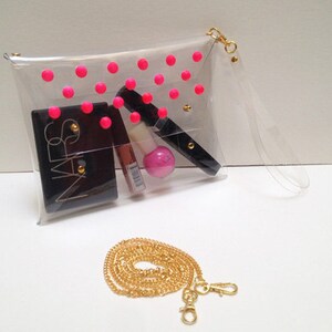 Puede incluir: Clutch transparente de pl&aacute;stico con un patr&oacute;n de lunares rosas y una correa de cadena dorada. La bolsa est&aacute; llena de art&iacute;culos de maquillaje, incluyendo un compacto negro NARS.
