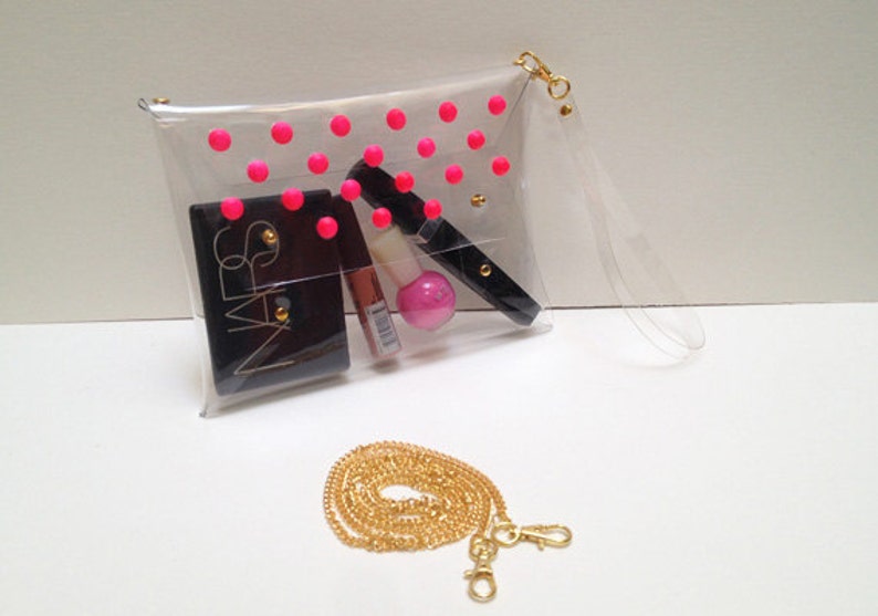 Puede incluir: Clutch transparente de pl&aacute;stico con un patr&oacute;n de lunares rosas y una correa de cadena dorada. La bolsa est&aacute; llena de art&iacute;culos de maquillaje, incluyendo un compacto negro NARS.