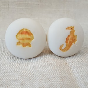 May include: Two white ceramic knobs with a seashell and a seahorse design. The seashell is yellow and orange with a white outline. The seahorse is yellow and orange with a white outline.