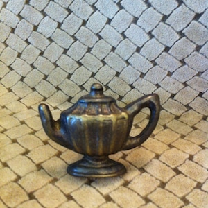 May include: A small, antique-looking, brass teapot with a fluted body and a handle on the side. The teapot is sitting on a patterned surface.