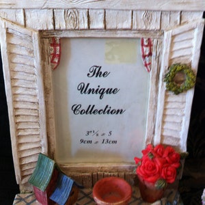 May include: White wooden picture frame with a cottage design. The frame has a red and white gingham ribbon, a green wreath, and a red flower pot with red roses. The frame has a white mat with the text "The Unique Collection 3 1/2 x 5 9cm x 13cm".