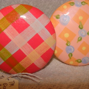 May include: Two round, painted, decorative ornaments. One is pink with a white and yellow plaid pattern. The other is pink with a white and yellow diamond pattern with blue flowers.