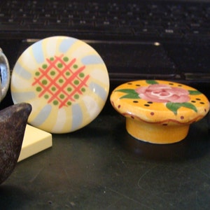 May include: A collection of five ceramic decorative knobs with various designs. The knobs feature a bird, a teapot, a floral pattern, a geometric pattern, and a floral design with a pink rose. The knobs are painted in various colors, including yellow, blue, green, red, and pink.