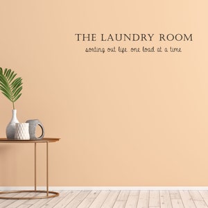 May include: A peach-colored wall with the text "THE LAUNDRY ROOM sorting out life one load at a time" on it. A gold metal side table with a vase and two decorative items on it.