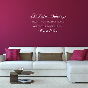 May include: A white wall with a pink wall decal that reads "A Perfect Marriage is just two imperfect people who refuse to give up on Each Other". A white sectional sofa with brown and pink throw pillows sits in front of the wall.