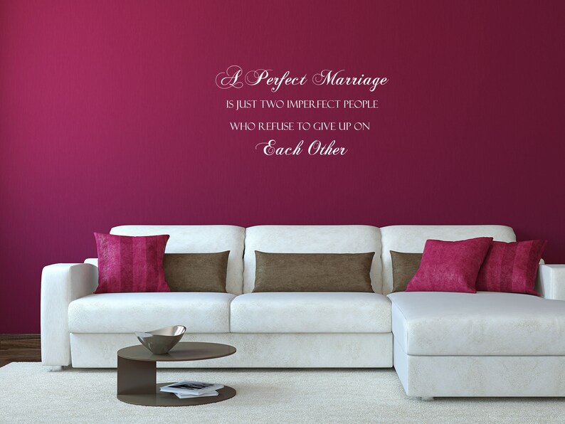 May include: A white wall with a pink wall decal that reads "A Perfect Marriage is just two imperfect people who refuse to give up on Each Other". A white sectional sofa with brown and pink throw pillows sits in front of the wall.